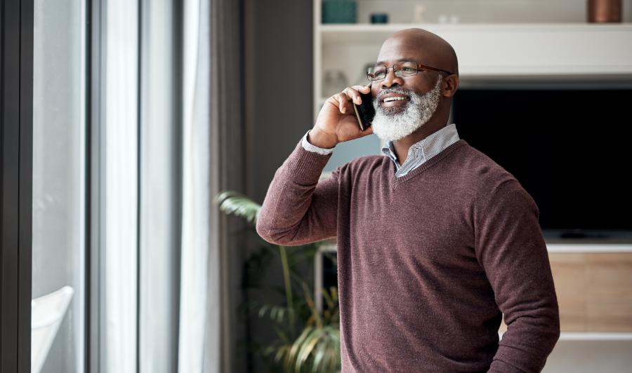 Older Black man smiles and stands at window while talking on the phone